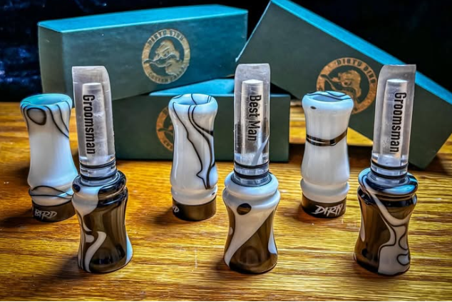 Five glass duck call lures with white, black, and gray marbled patterns are arranged on a wooden surface, with two teal boxes and a round sticker featuring the 'Alty Bird Custom Calls' logo in the background.